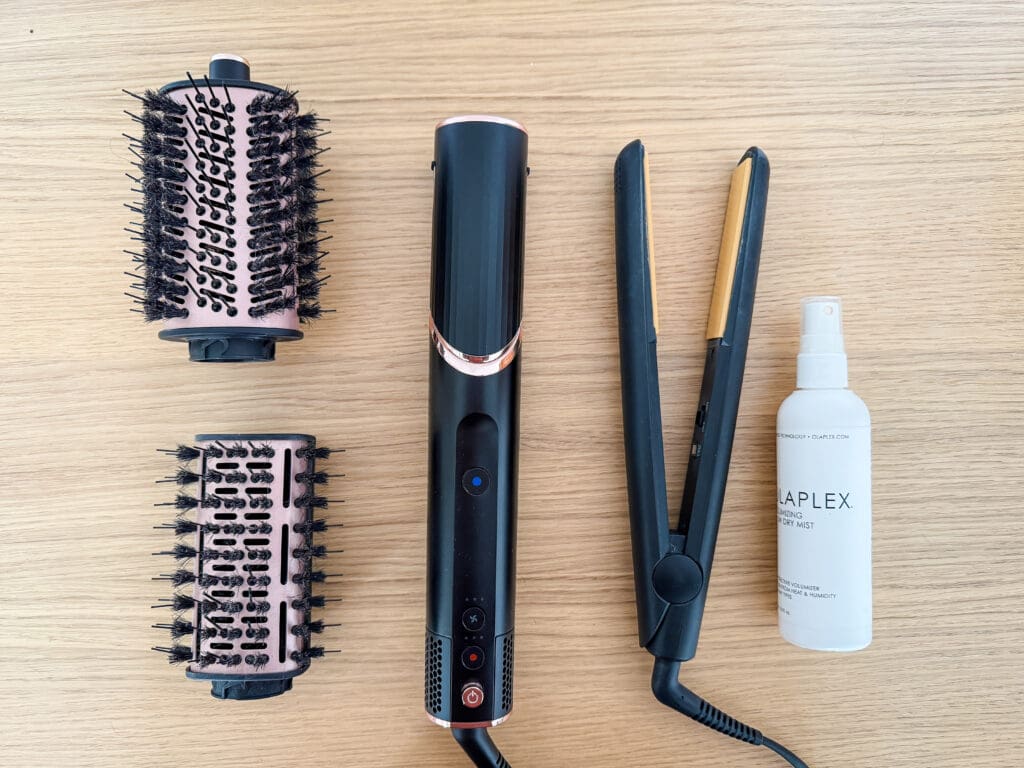 A flat lay comparison of hair tools on a wooden desk, featuring the Shark FlexStyle base with two brush attachments, a classic GHD flat iron, and a bottle of Olaplex Volumizing Blow Dry Mist.