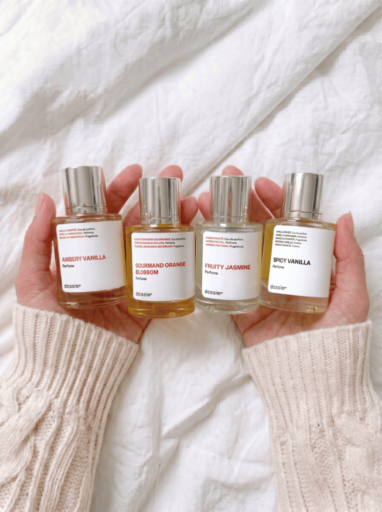 A person wearing a cream cable-knit sweater holding four Dossier perfume bottles: Ambery Vanilla, Gourmand Orange Blossom, Fruity Jasmine, and Spicy Vanilla. The bottles are lined up in their hands against a soft white linen background.