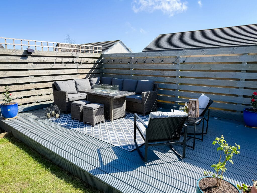 A full view of a grey composite deck with a large rattan corner sofa, fire pit table, and blue patterned outdoor rug.