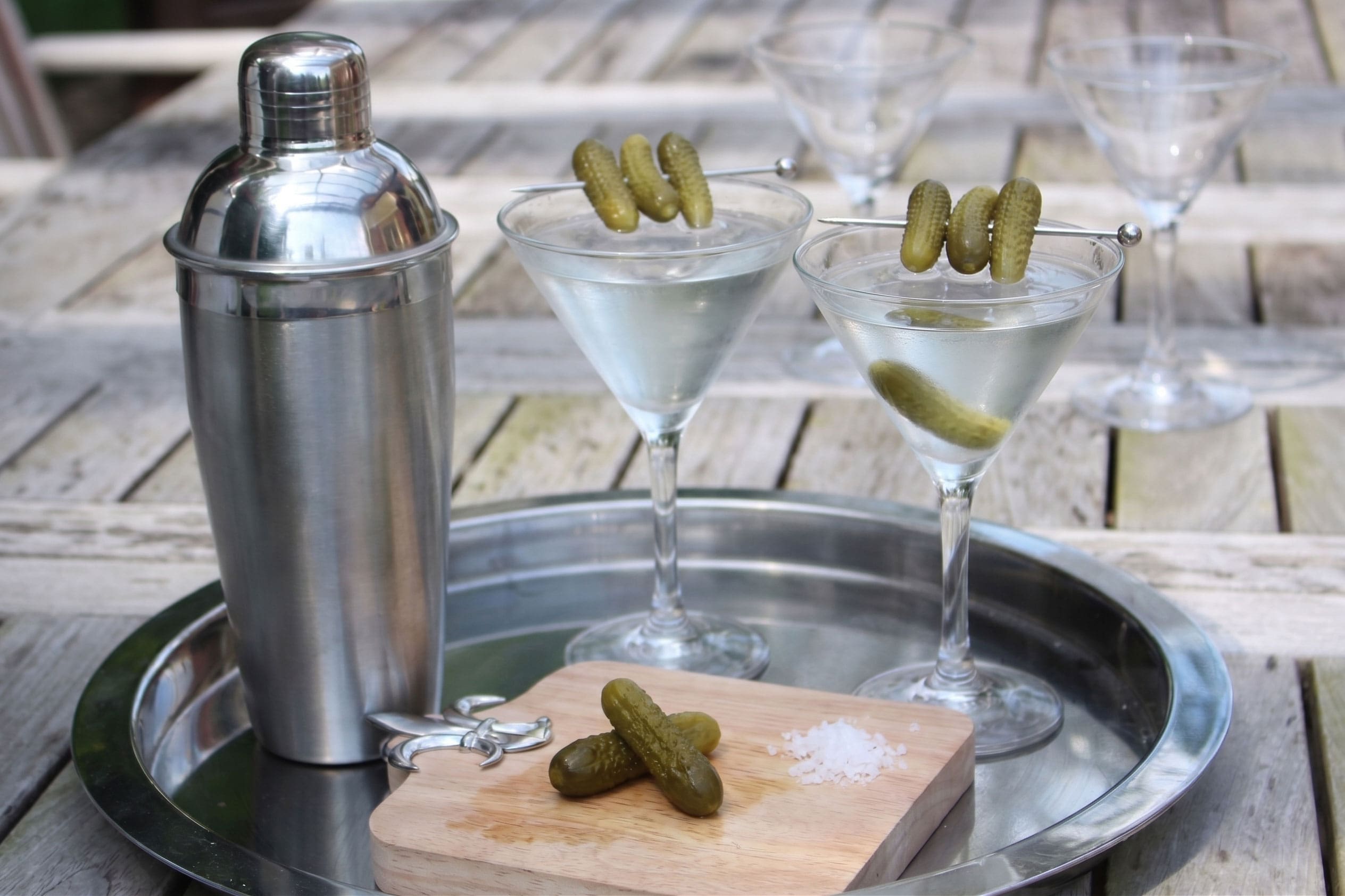 Pickle martini in a chilled glass beside a cocktail shaker on a sunny outdoor table, garnished with a gherkin