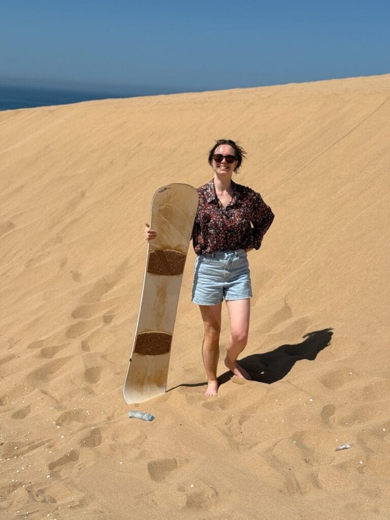 Woman in a floral shirt and denim shorts holding a sandboard on a golden dune near Agadir