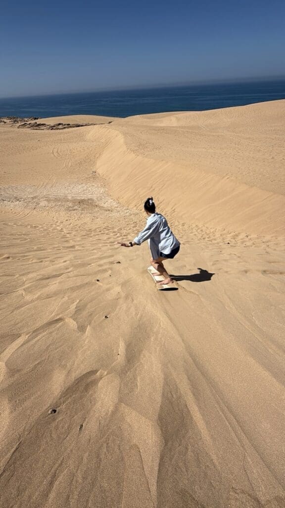 Woman sandboarding down a golden dune with the Atlantic Ocean in the background