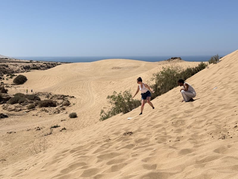 Two women climbing a steep sand dune after sandboarding near Agadir